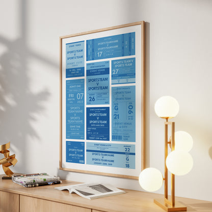 A framed poster featuring blue and white sports team event tickets, displayed on a wooden desk in a modern room setting next to a multi-light lamp.