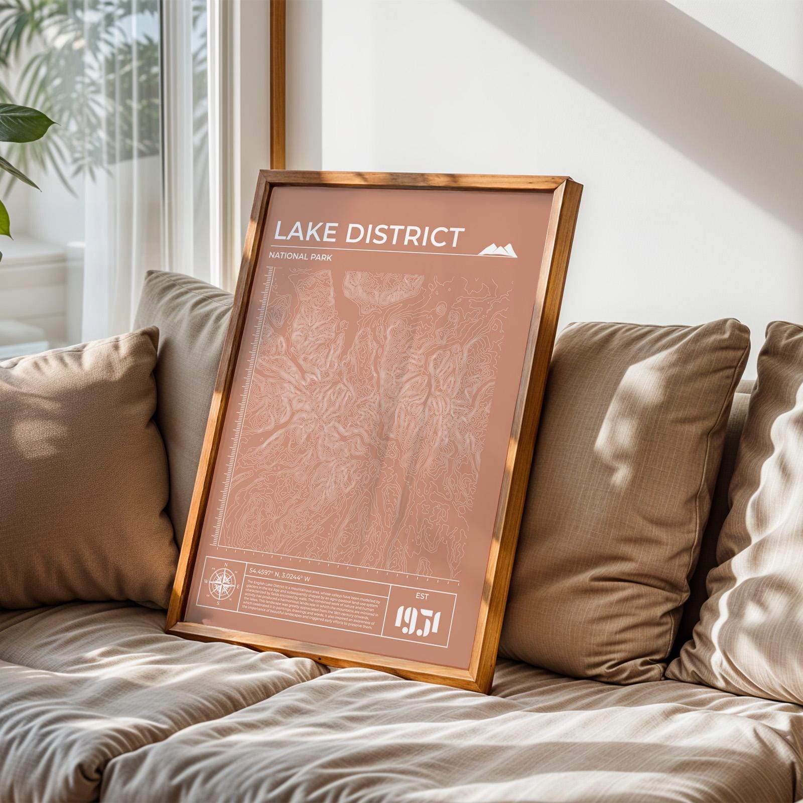 Framed Lake District map art poster in terracotta on sofa, perfect gift for hikers and outdoor adventure lovers.