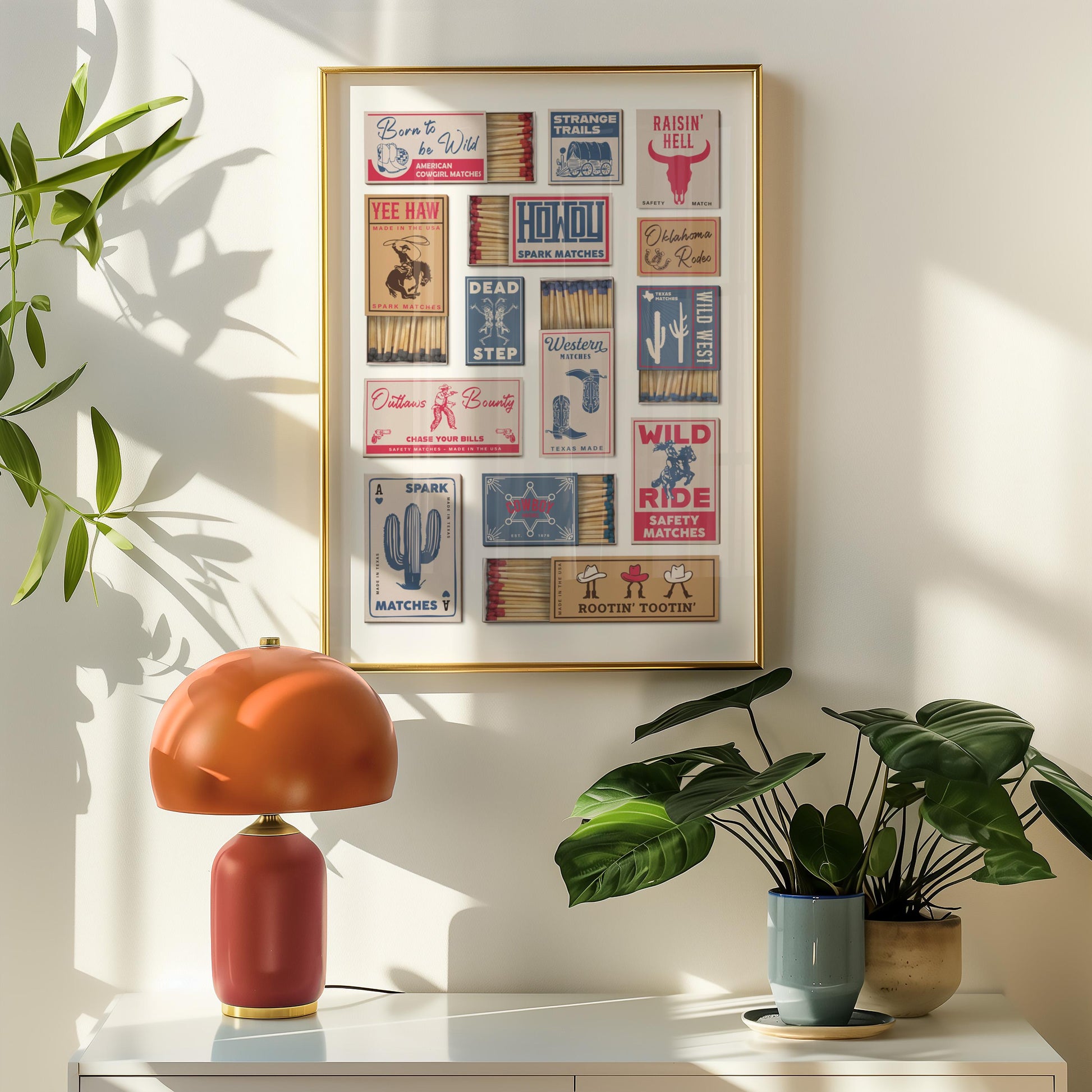 Eclectic home decor with vintage matchbox labels, featuring cowboy-themed designs and text such as "Outlaw's Bounty". The artwork hangs above a red and orange table lamp and potted plants, creating a stylish interior.