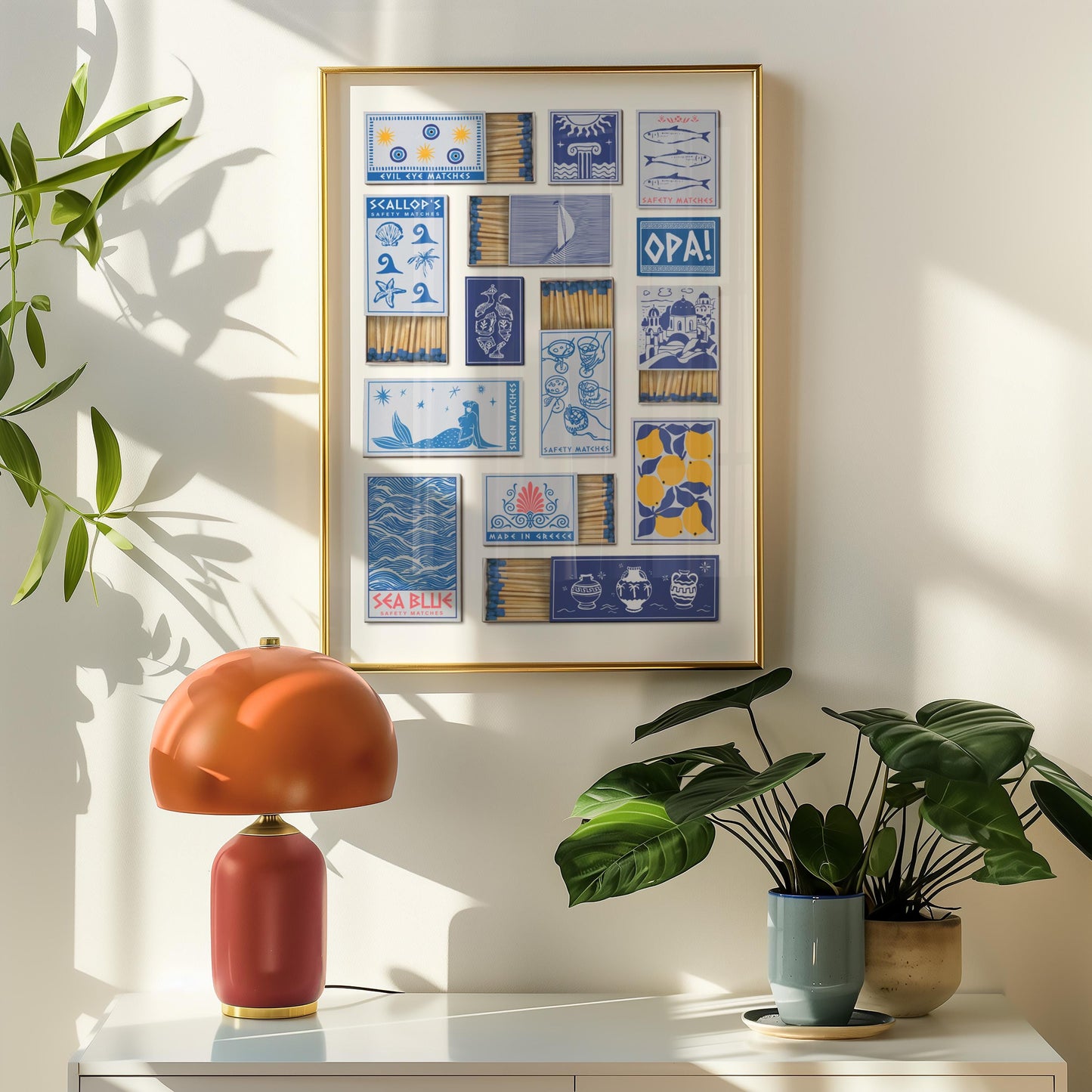A home decor piece displaying blue and yellow matchbox designs, with text including "Made In Greece"." Below, a red and orange table lamp with a gold base. Green plants in pots are on the right, creating a balanced composition.