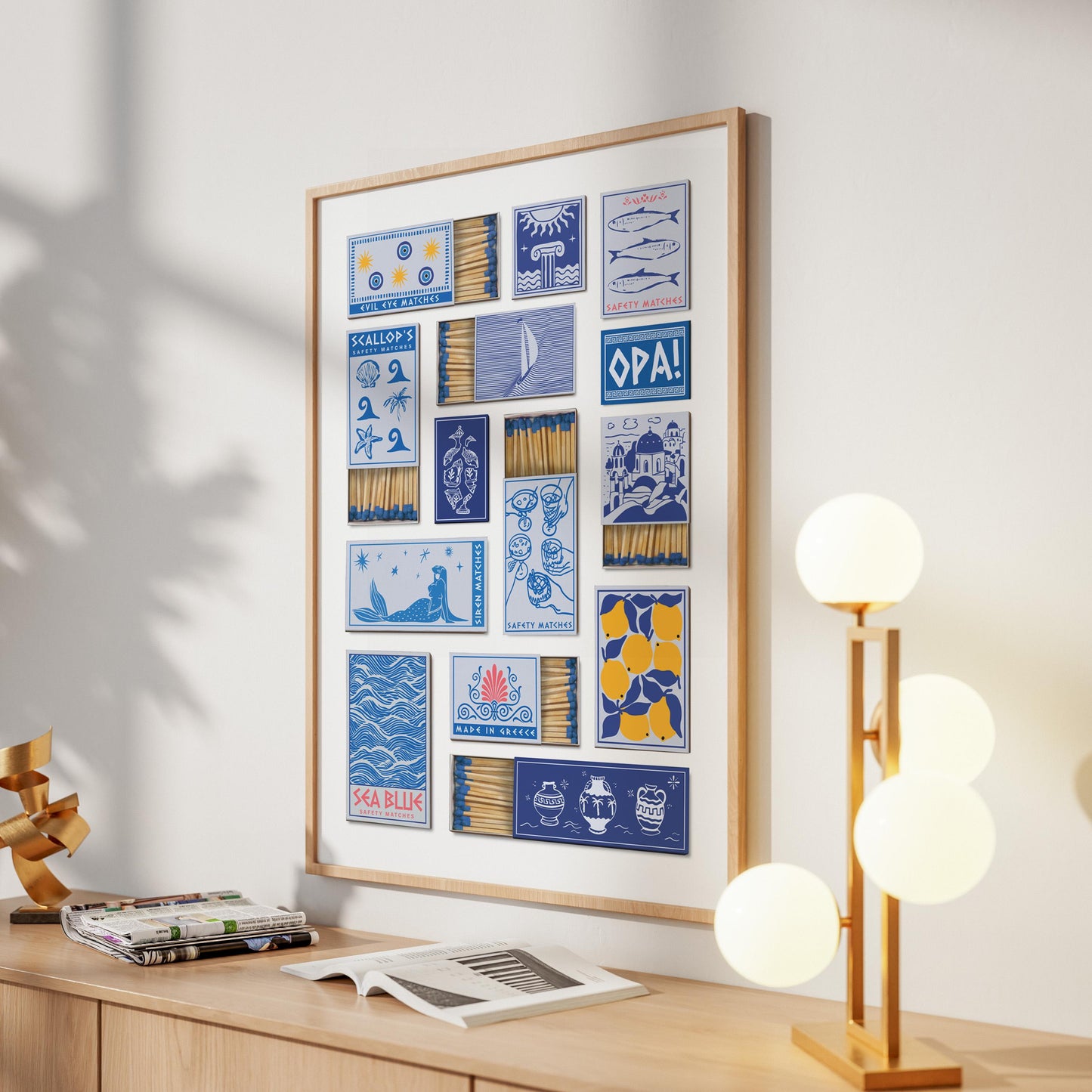 A print in a frame displaying a collection of matchbook designs. The designs include images of fish, a sailboat and the word "OPA!". The frame is light brown, and the print is on a white wall.