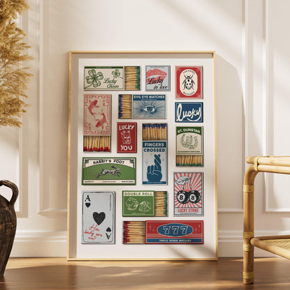 A framed print displaying vintage matchbox designs with lucky symbols and phrases. The artwork includes images of a four-leaf clover and playing card, with text like "Fingers Crossed". The frame is gold.