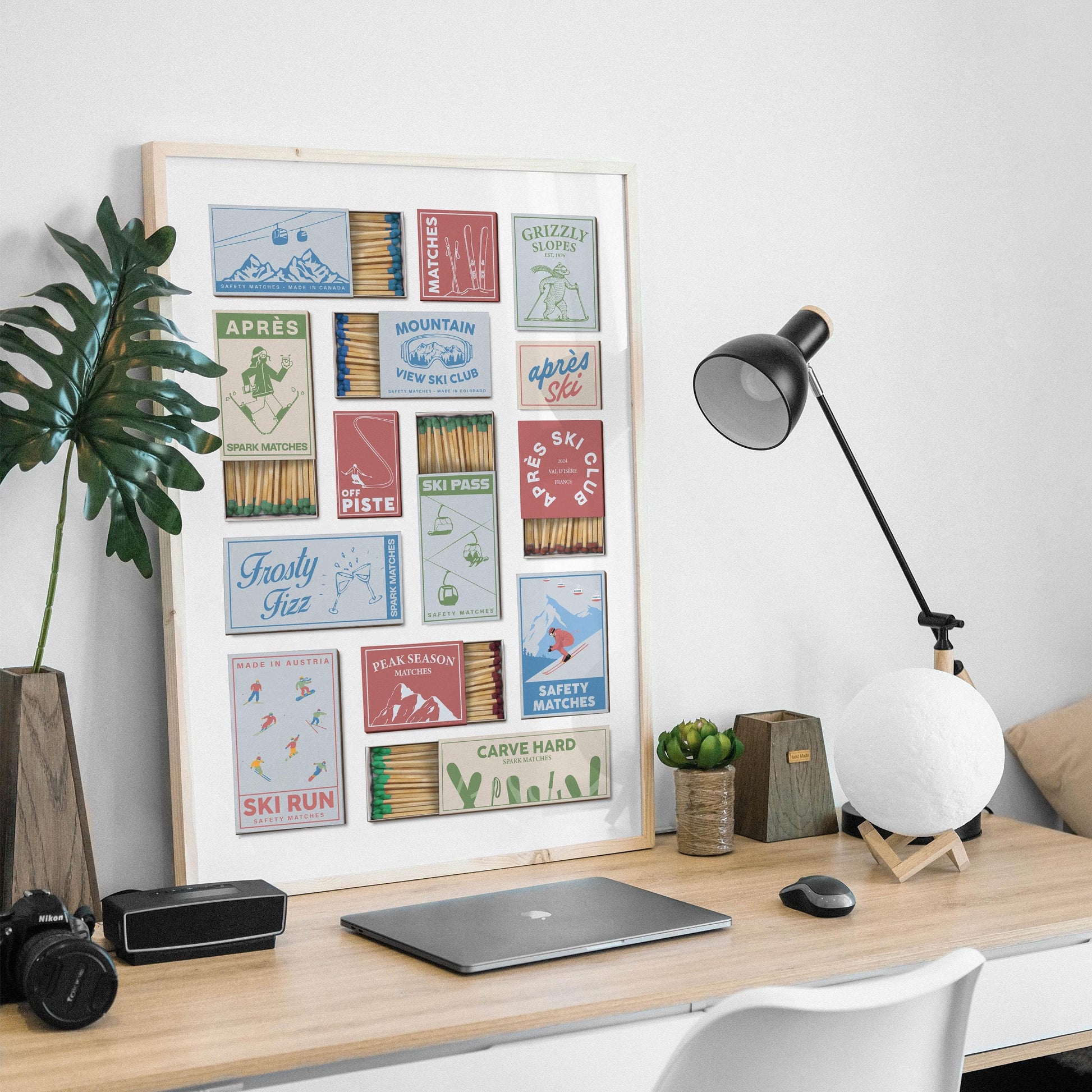 A print in a frame showcasing vintage matchbox designs with ski-themed illustrations and text. The matchbook designs are in various colours, including red, blue, and green, with phrases like "Après Ski". The print is displayed on a wooden desk.