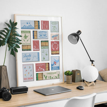 A print in a frame showcasing vintage matchbox designs with ski-themed illustrations and text. The matchbook designs are in various colours, including red, blue, and green, with phrases like "Après Ski". The print is displayed on a wooden desk.