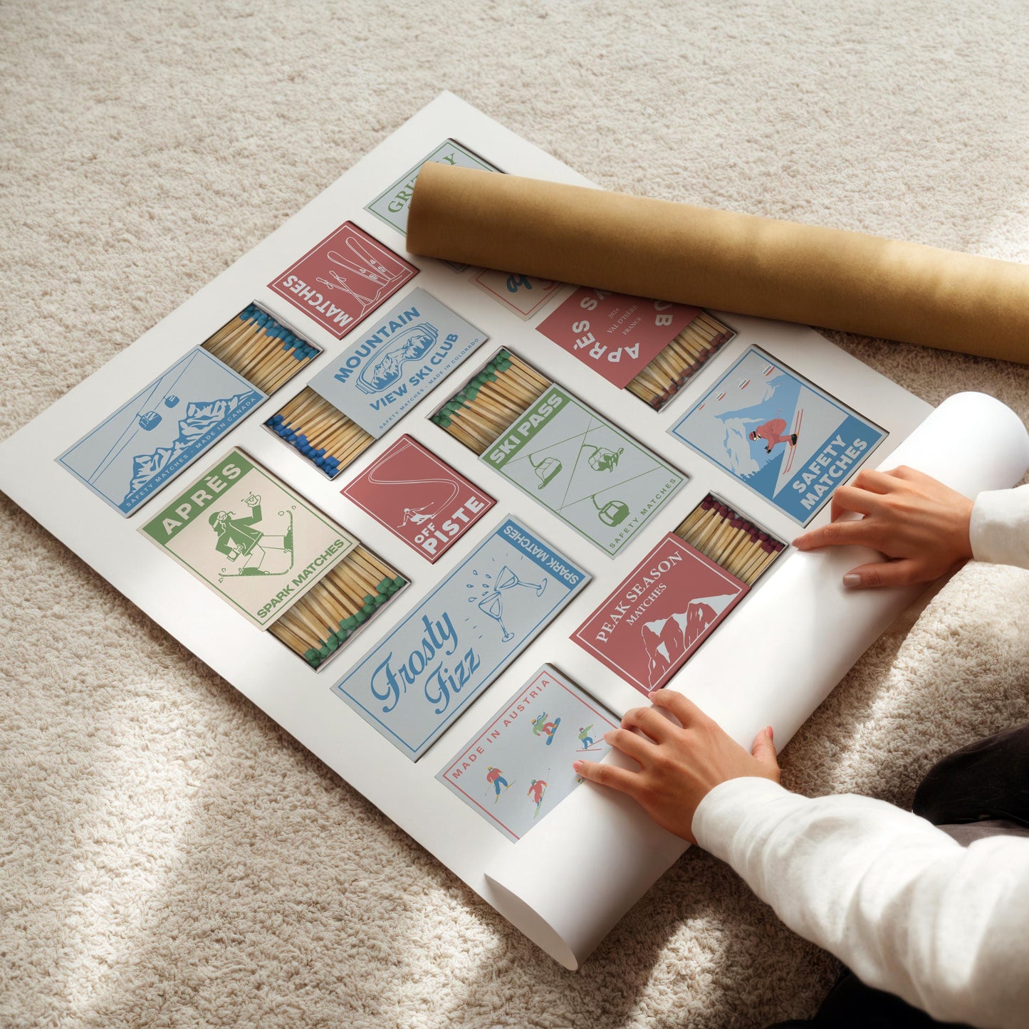 A poster with vintage-style matchbox designs, themed around skiing resorts. The matchboxes are in various colours and feature illustrations of skiers, ski lifts, and text such as "Ski Pass". A rolled-up poster is next to the poster.