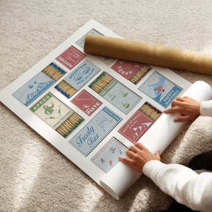 A poster with vintage-style matchbox designs, themed around skiing resorts. The matchboxes are in various colours and feature illustrations of skiers, ski lifts, and text such as "Ski Pass". A rolled-up poster is next to the poster.