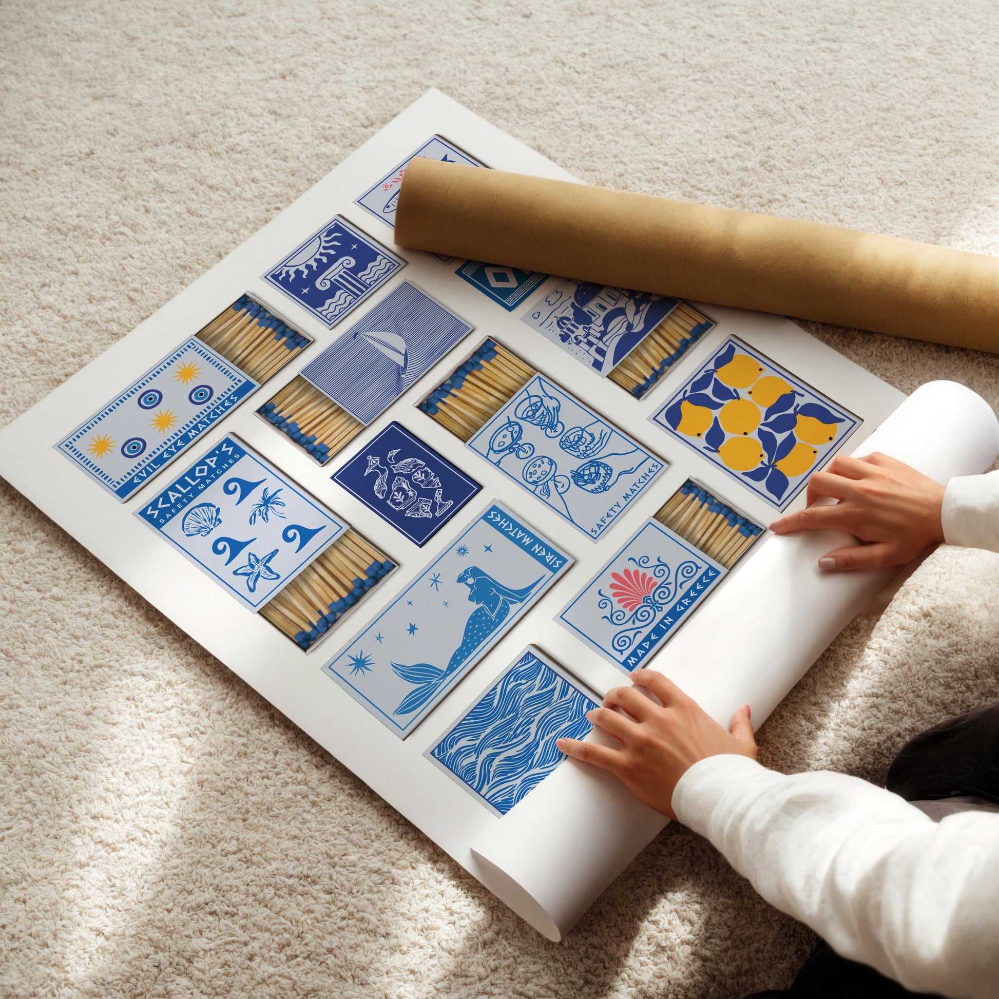 A large white poster with blue and yellow illustrated matchbook designs, including a statue, scallops, and lemons. A rolled-up poster is next to the main poster. The text "Siren Matches" and "evil eye" are visible.