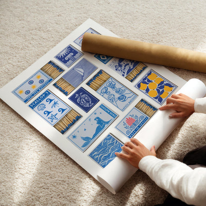 A large white poster with blue and yellow illustrated matchbook designs, including a statue, scallops, and lemons. A rolled-up poster is next to the main poster. The text "Siren Matches" and "evil eye" are visible.