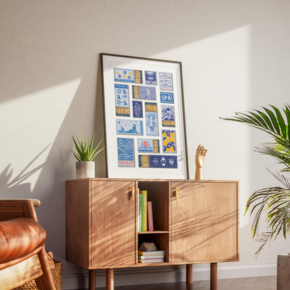 A trendy downloadable art print displaying various illustrated greek matchbooks designs in blue, yellow, and white. The print is on a wooden cabinet with gold handles, accompanied by a potted plant and a wooden hand sculpture.