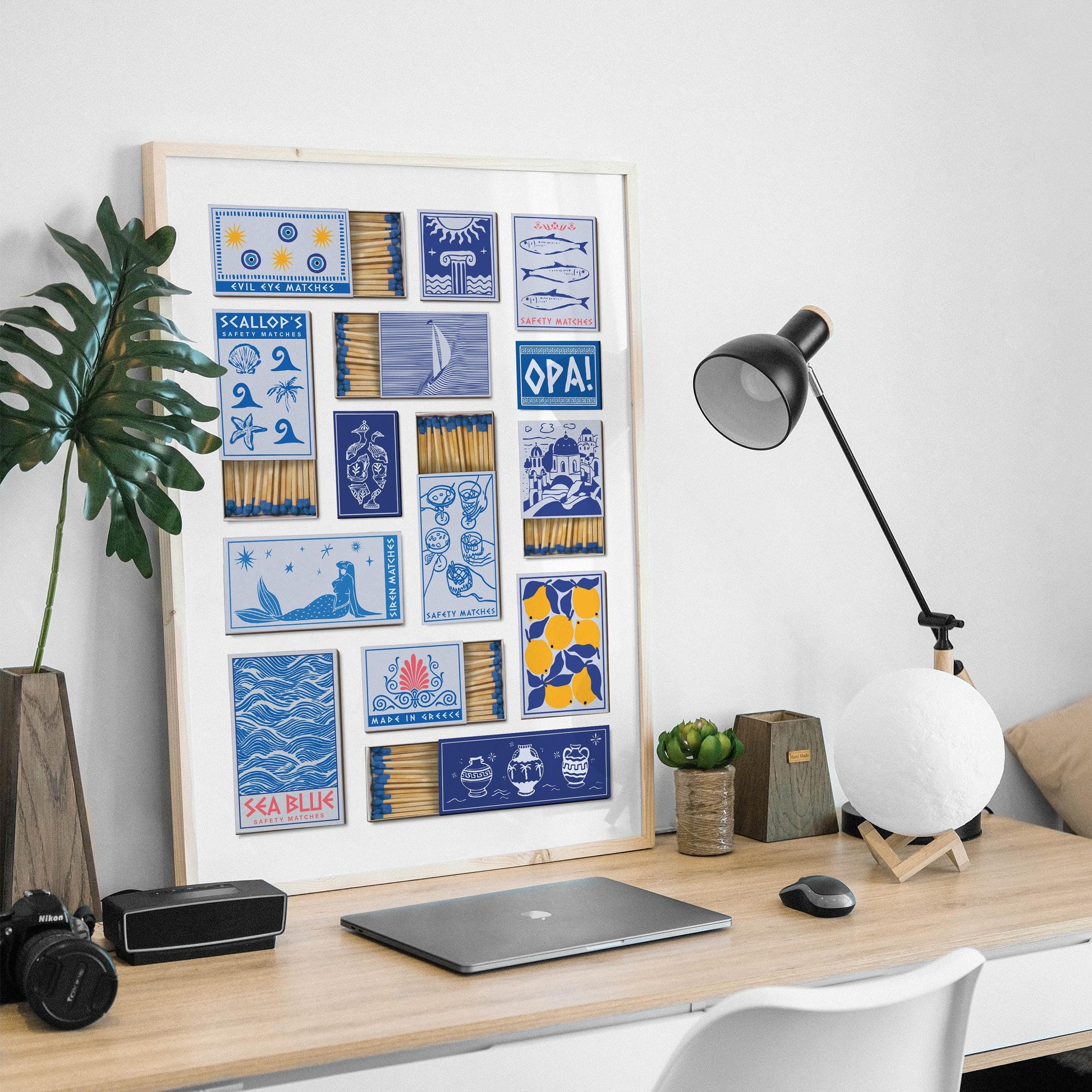 A framed art printable decor option with blue and yellow matchbox designs. The designs include illustrations of lemons, blue domes, and the text "OPA!". The artwork is displayed on a wooden desk with a lamp and laptop in a mans office in an apartment.