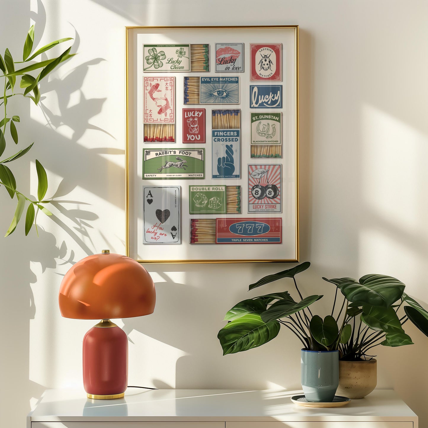 A wall art piece displaying vintage matchbox designs with luck symbols and text. The artwork hangs above a white surface, with a red and orange table lamp and potted plants in the foreground.