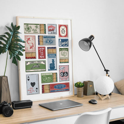 A framed print displaying vintage matchbox designs with themes of luck and fortune. The matchbooks are in various colours and feature text including "Lucky in Love". The print is on a wooden desk, with a lamp and laptop in a mans office in an apartment.