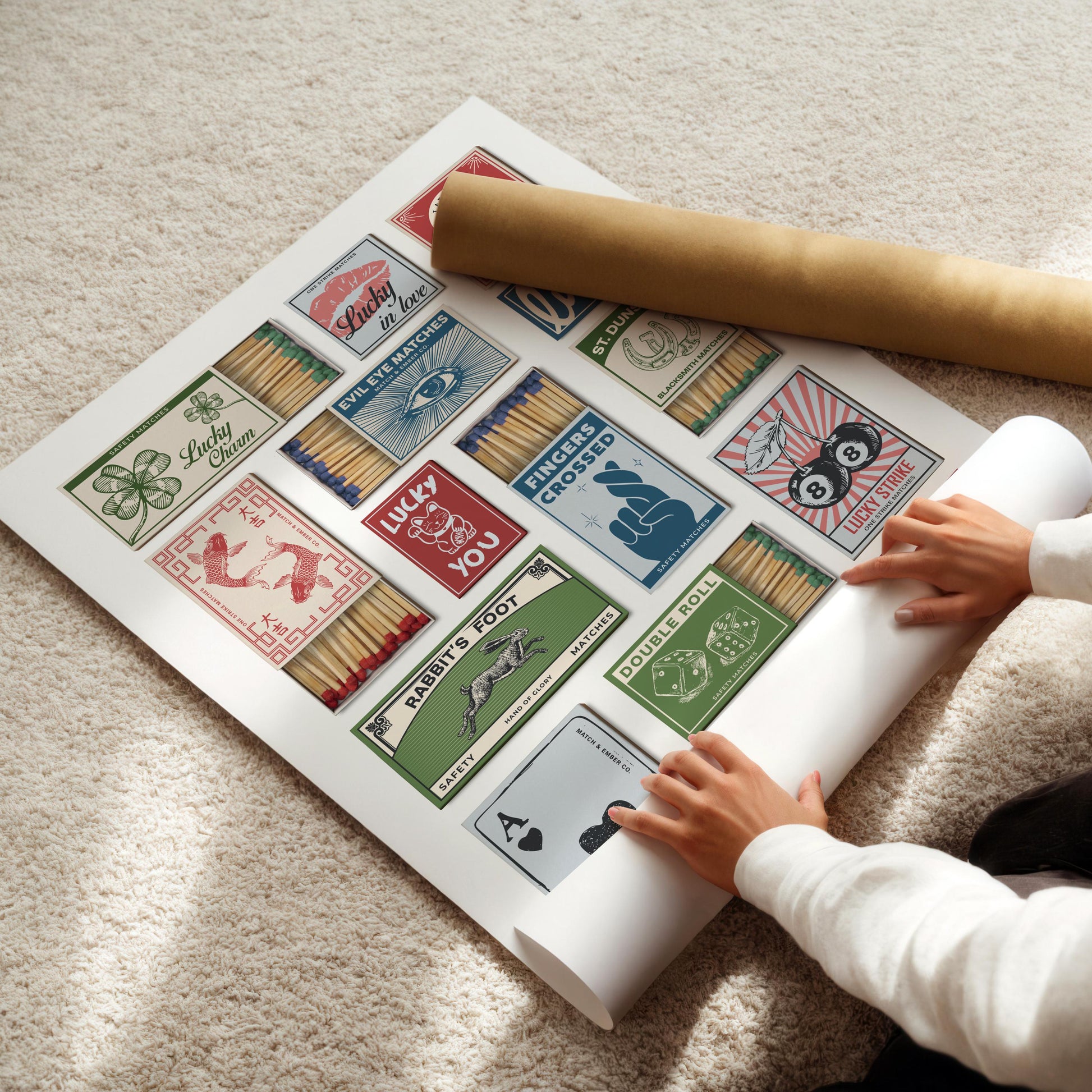 A funky art piece with multiple matchbox designs, each with a different lucky theme. The designs include text such as "Lucky Charm," "Fingers Crossed," and "Rabbit's Foot." A rolled poster and hands are visible.