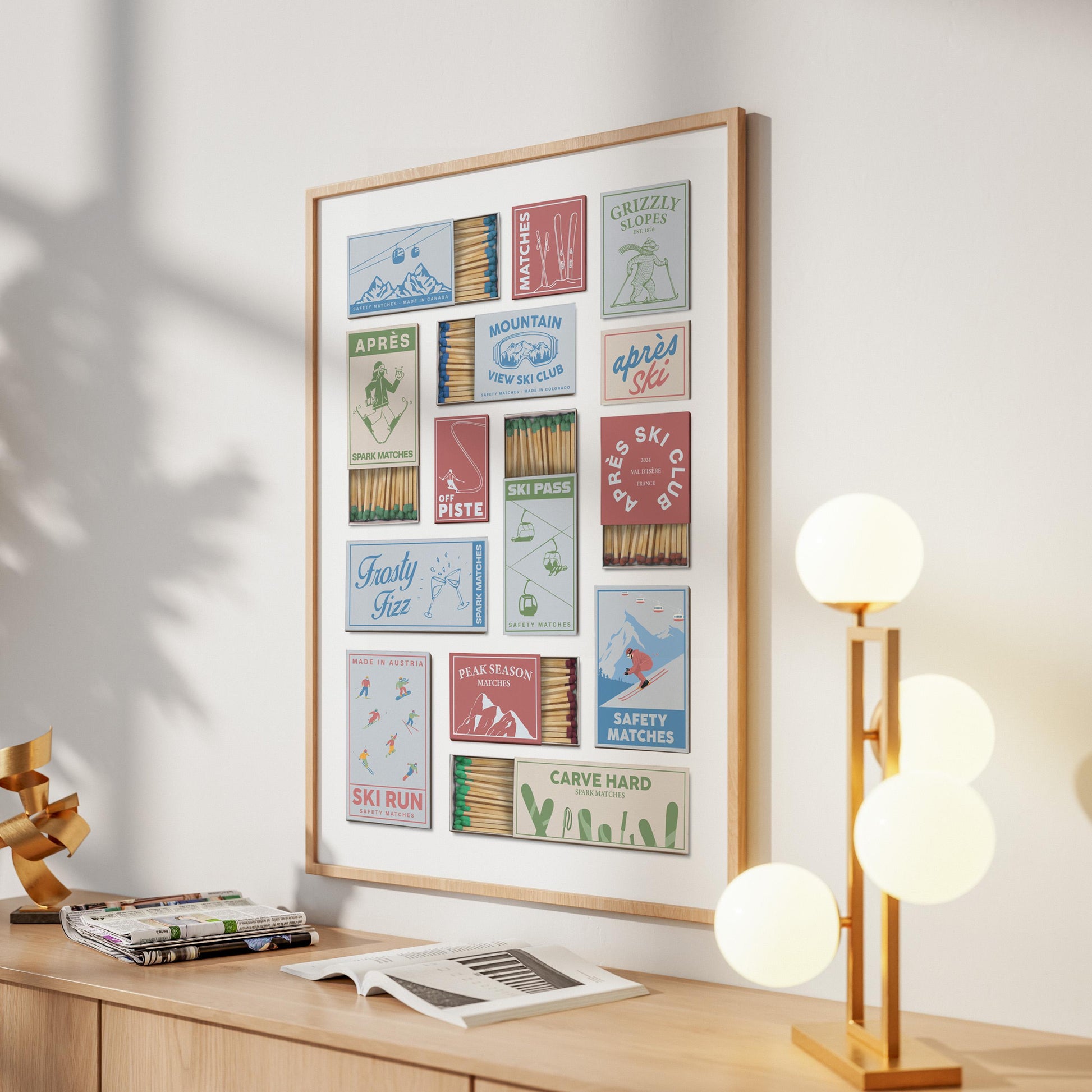 A framed print showcasing vintage matchbook designs with snowboard resort themes. The boxes are in various colours and feature text such as "Après Ski" and "Grizzly Slopes". The artwork is displayed on a light-coloured wall.