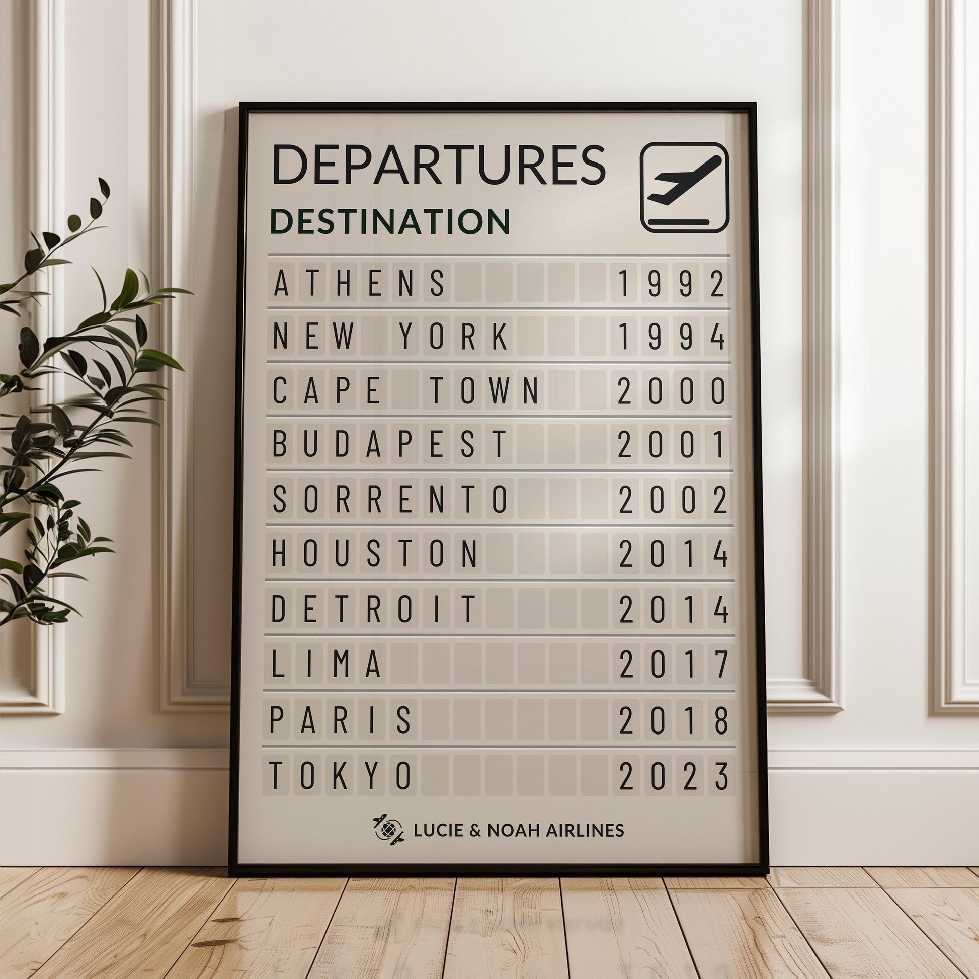A minimalist home with white cladded walls and a black framed print leaning on the wall. The print is inspired by departure boards in the airport featuring places like Athens, Paris and Tokyo with dates from the holiday.