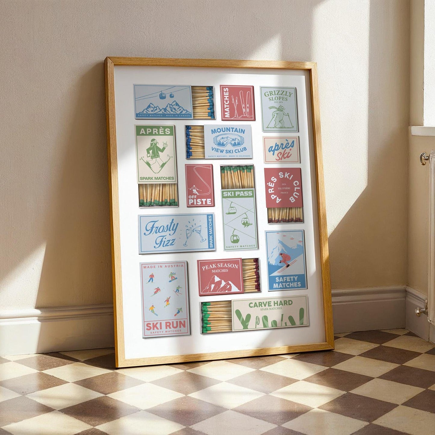 A framed print with a collection of vintage-style matchbox designs. The boxes feature ski-themed illustrations and text, including skiers, mountains, and resort names. The frame is a light brown colour, and the print is displayed indoors.