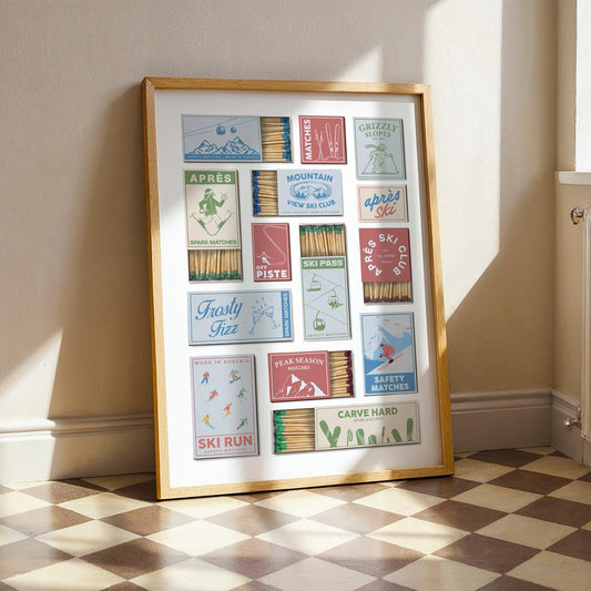 A framed print with a collection of vintage-style matchbox designs. The boxes feature ski-themed illustrations and text, including skiers, mountains, and resort names. The frame is a light brown colour, and the print is displayed indoors.