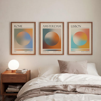 Three framed art prints, each displaying a city name: Rome, Amsterdam, and Lisbon. The gallery wall prints showcase a gradient aura design in various colours. They are hung in a bedroom, with a wooden bedside table and a lamp.