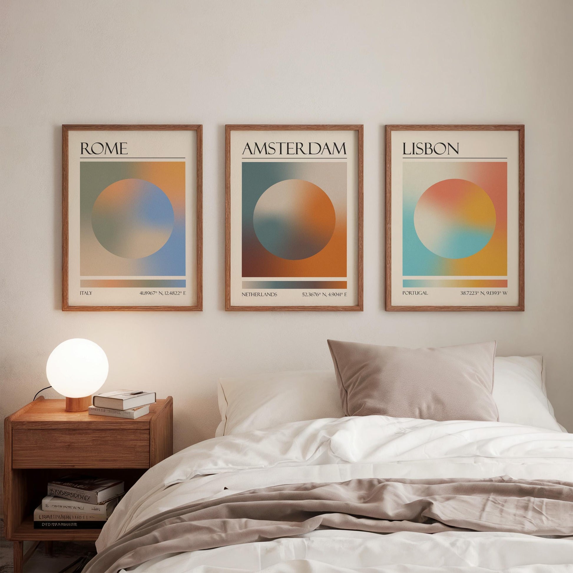Three framed art prints, each displaying a city name: Rome, Amsterdam, and Lisbon. The gallery wall prints showcase a gradient aura design in various colours. They are hung in a bedroom, with a wooden bedside table and a lamp.