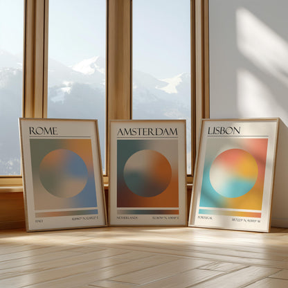 Three framed art prints with abstract designs and gradient colour palettes. The set of three triptych prints each display a city name: Rome, Amsterdam, and Lisbon. The prints are in neutral-toned wooden frames, leaning against a window.