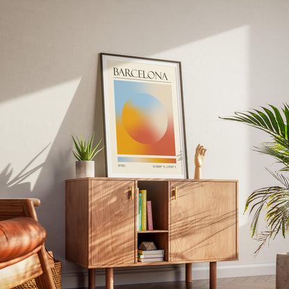 A black frame with an aura gradient circle print inside. The word "BARCELONA" and coordinates are at the bottom. The memory keepsake print is displayed on a wooden cabinet with gold handles.