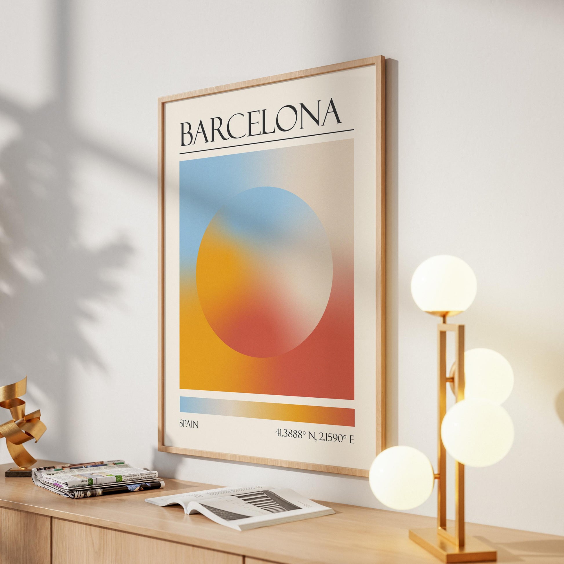 A framed print with the word "BARCELONA" at the top. The destination artwork features a colourful aura circle in teal, orange, and red. A gold lamp with four white globe lights is to the right in the hallway.