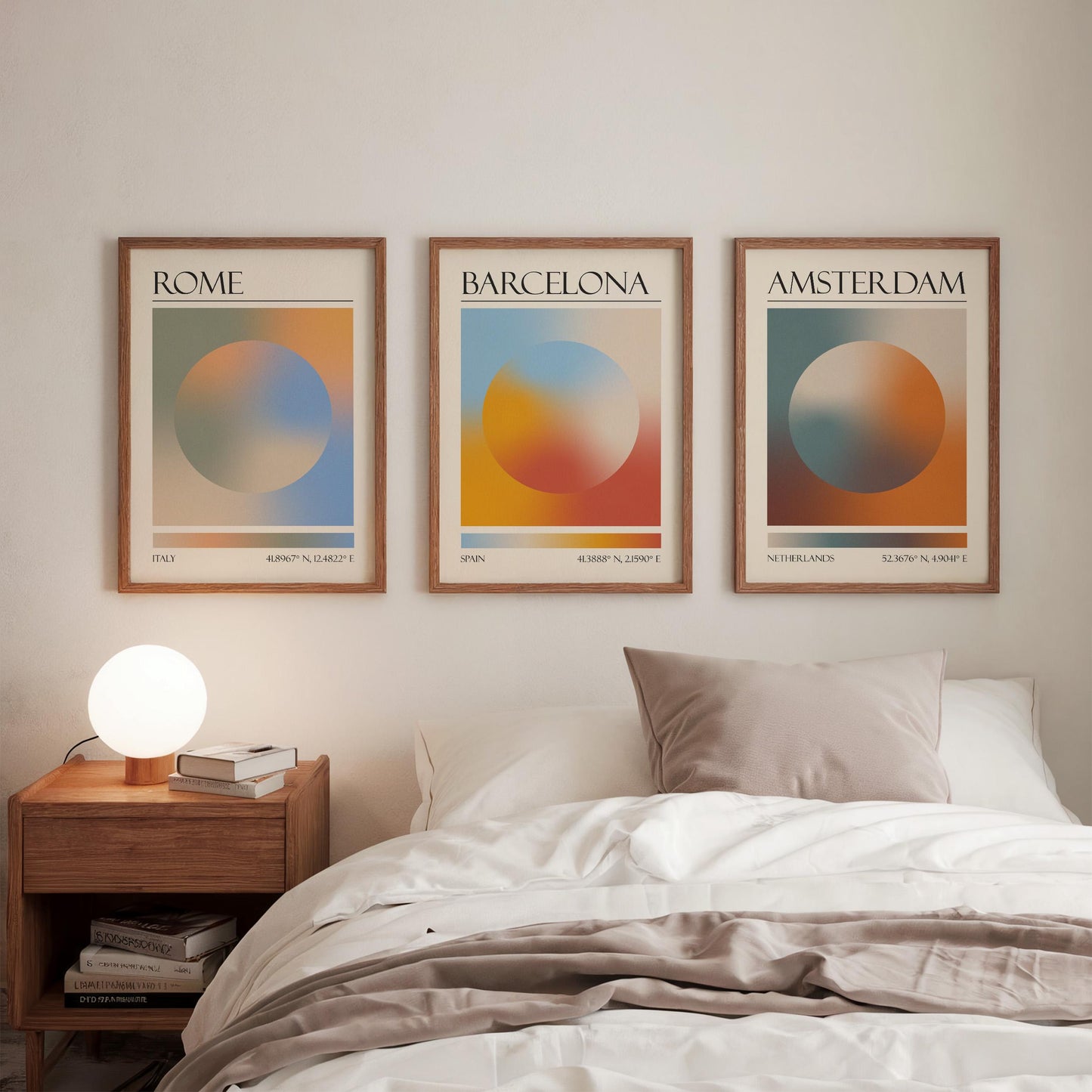 Three framed art prints, each displaying a city name: Rome, Amsterdam, and Barcelona. The gallery wall prints showcase a gradient aura design in various colours. They are hung in a bedroom, with a wooden bedside table and a lamp.
