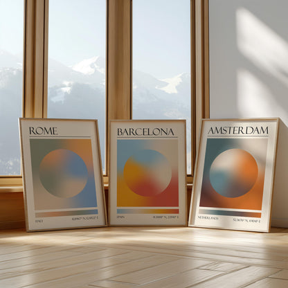 Three framed art prints with abstract designs and gradient colour palettes. The set of three triptych prints each display a city name: Rome, Amsterdam, and Barcelona. The prints are in neutral-toned wooden frames, leaning against a window.