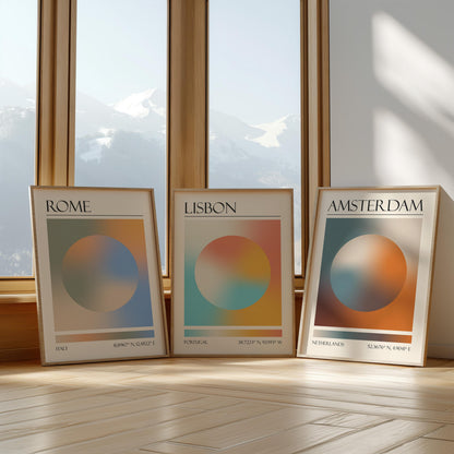 Three framed art prints with abstract designs and gradient colour palettes. The set of three triptych prints each display a city name: Rome, Amsterdam, and Lisbon. The prints are in neutral-toned wooden frames, leaning against a window.