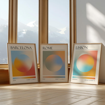 Three framed art prints with abstract designs and gradient colour palettes. The set of three triptych prints each display a city name: Rome, Barcelona, and Lisbon. The prints are in neutral-toned wooden frames, leaning against a window.