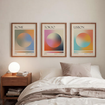 Three framed art prints, each displaying a city name: Rome, Tokyo, and Lisbon. The gallery wall prints showcase a gradient aura design in various colours. They are hung in a bedroom, with a wooden bedside table and a lamp.