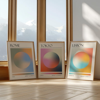 Three framed art prints with abstract designs and gradient colour palettes. The set of three triptych prints each display a city name: Rome, Tokyo, and Lisbon. The prints are in neutral-toned wooden frames, leaning against a window.