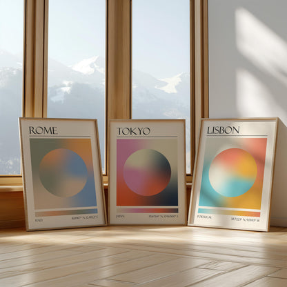 Three framed art prints with abstract designs and gradient colour palettes. The set of three triptych prints each display a city name: Rome, Tokyo, and Lisbon. The prints are in neutral-toned wooden frames, leaning against a window.