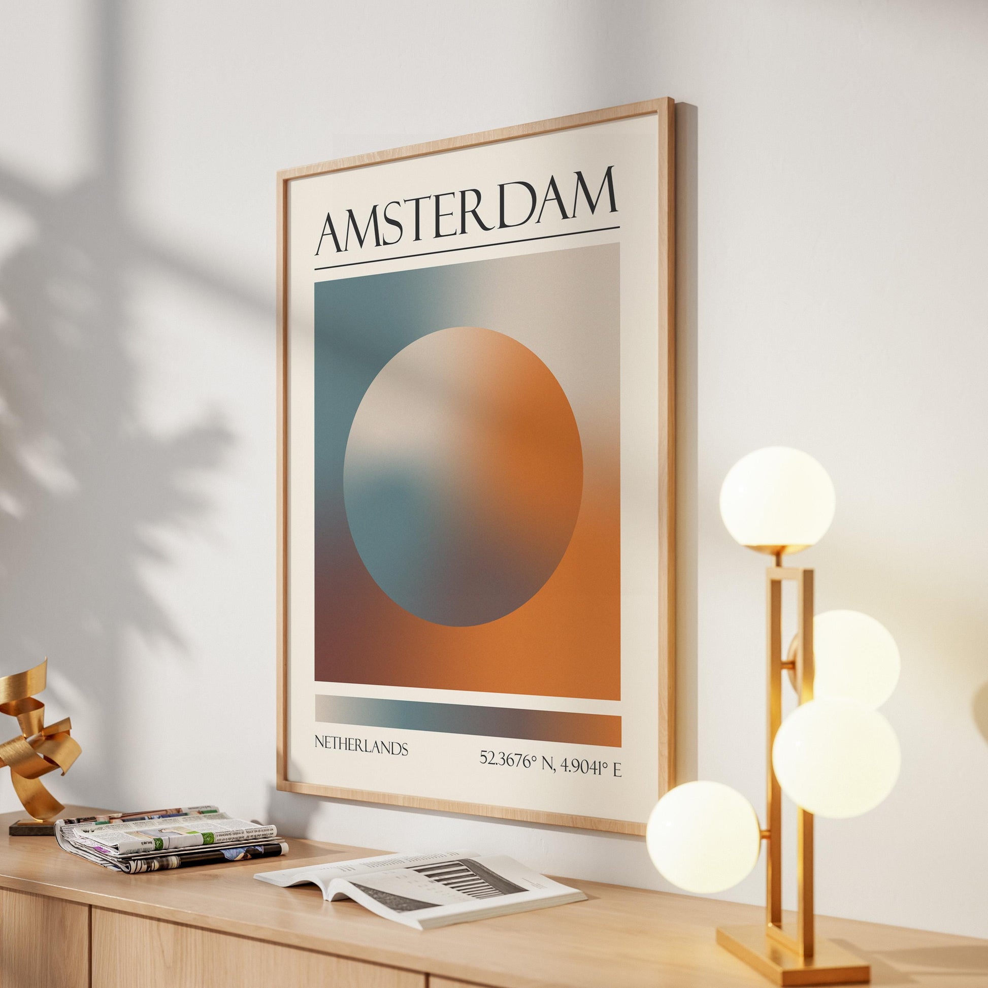 A framed print with the word "AMSTERDAM" at the top. The destination artwork features a colourful aura circle in teal, orange, and brown. A gold lamp with four white globe lights is to the right in the hallway.