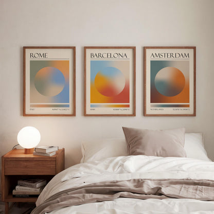Three framed art prints, each displaying a city name: Rome, Amsterdam, and Barcelona. The gallery wall prints showcase a gradient aura design in various colours. They are hung in a bedroom, with a wooden bedside table and a lamp.