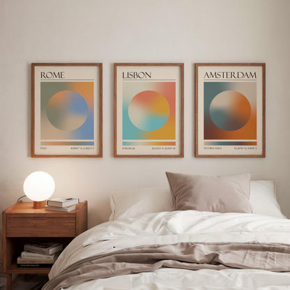 Three framed art prints, each displaying a city name: Rome, Amsterdam, and Lisbon. The gallery wall prints showcase a gradient aura design in various colours. They are hung in a bedroom, with a wooden bedside table and a lamp.