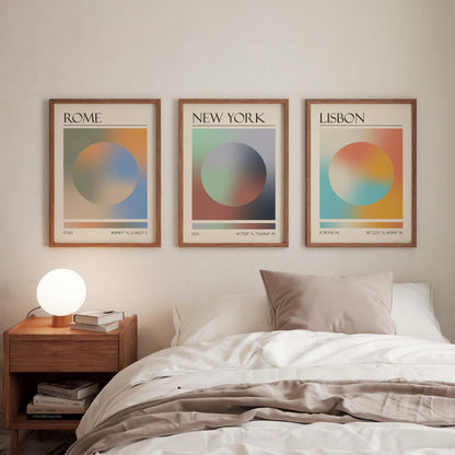 Three framed art prints, each displaying a city name: Rome, New York, and Lisbon. The gallery wall prints showcase a gradient aura design in various colours. They are hung in a bedroom, with a wooden bedside table and a lamp.