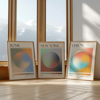 Three framed art prints with abstract designs and gradient colour palettes. The set of three triptych prints each display a city name: Rome, New York, and Lisbon. The prints are in neutral-toned wooden frames, leaning against a window.
