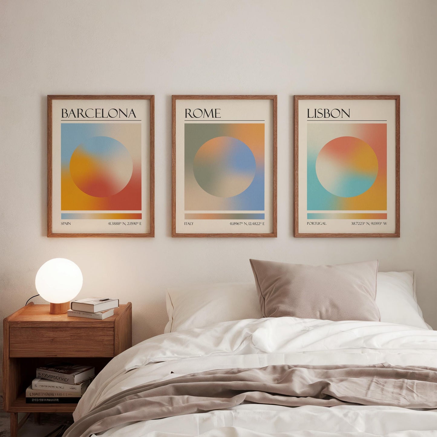 Three framed art prints, each displaying a city name: Rome, Barcelona, and Lisbon. The gallery wall prints showcase a gradient aura design in various colours. They are hung in a bedroom, with a wooden bedside table and a lamp.