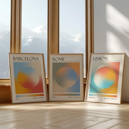 Three framed art prints with abstract designs and gradient colour palettes. The set of three triptych prints each display a city name: Rome, Barcelona, and Lisbon. The prints are in neutral-toned wooden frames, leaning against a window.