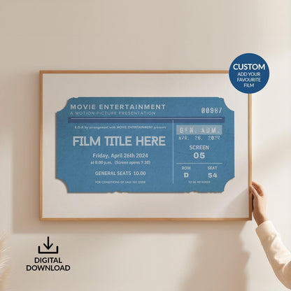 Someone holding up a large horizontal frame on a white wall. The art is a muted rich blue coloured movie ticket inspired by old movie theatre stubs