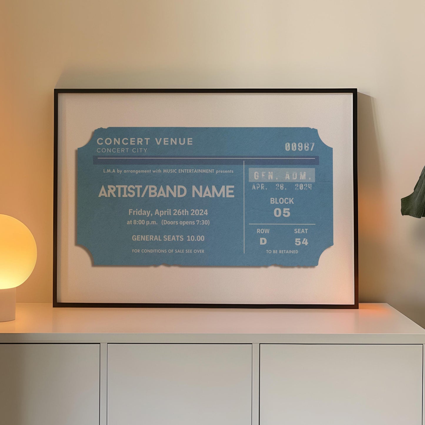 A blue coloured concert ticket design on a poster in a black frame in a warm lit office space. The art is personalised and has custom text details.