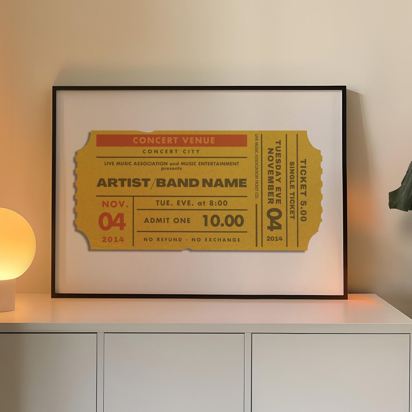 A yellow coloured concert ticket design on a poster in a black frame in a warm lit office space. The art is personalised and has custom text details.