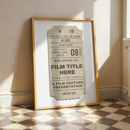 A framed vintage-style film ticket art print leaning against a beige wall on a checkered floor. The print has a cream ticket design with black text, set in a white mat and a thin gold frame.