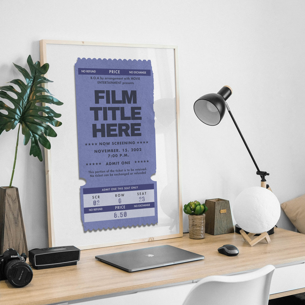 Framed movie ticket poster on a desk with a laptop, lamp, and plant.
